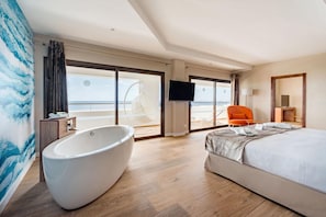 Een moderne hotelkamer met een groot bad, een bed, een televisie en uitzicht op het strand.