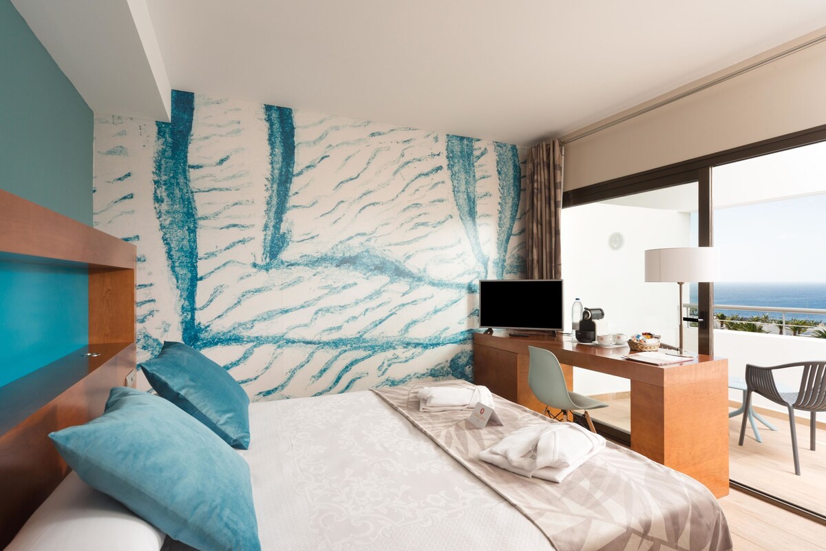 A hotel room with a bed, a desk, a chair, a television, and a view of the ocean.