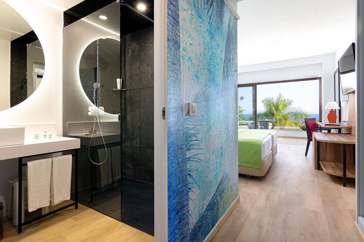 A modern bathroom with a large mirror, a shower, and a sink. There is a bed with a view of the outdoors.