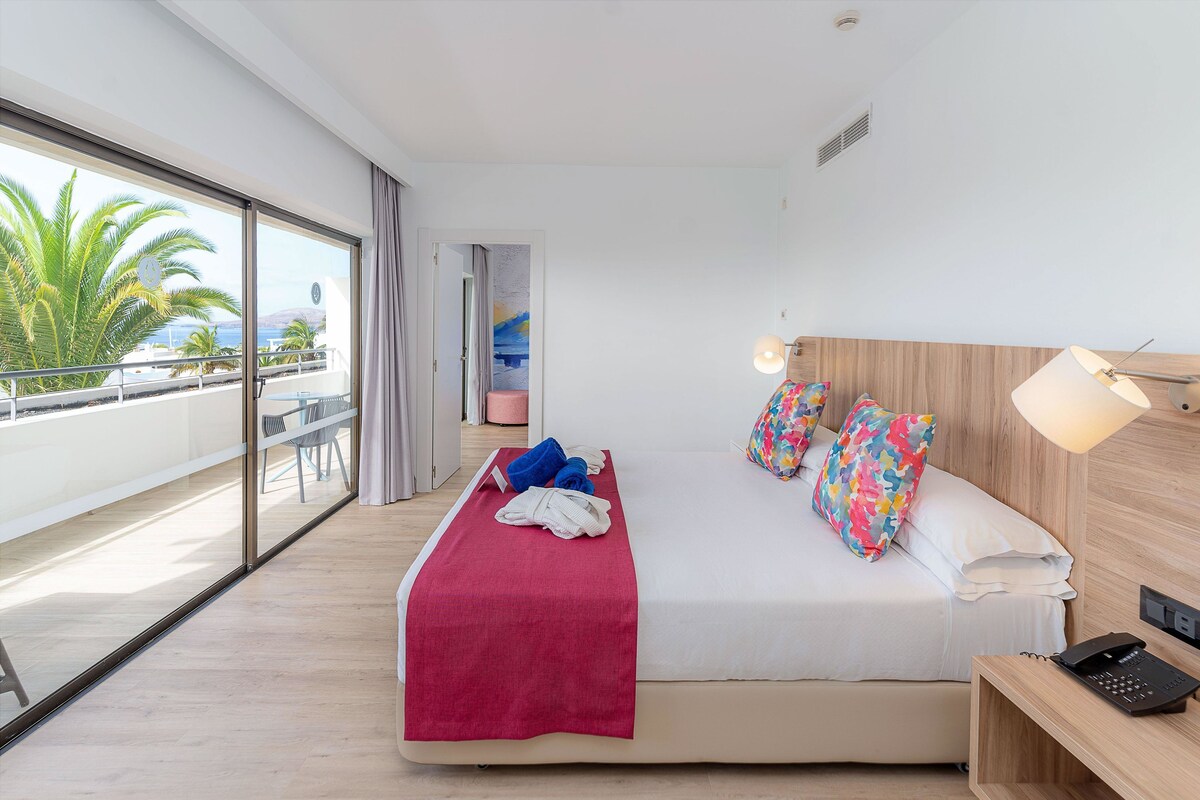 Une chambre d’hôtel moderne dotée d’un grand lit, d’une table de chevet avec une lampe, et offrant une vue sur les palmiers et la mer grâce à de grandes fenêtres.