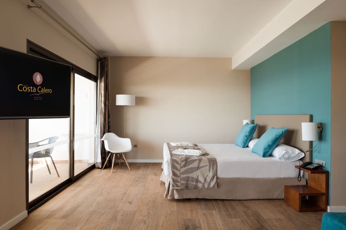Ein modernes Hotelzimmer mit Bett, Nachttisch, Lampe und Blick durch ein großes Fenster nach draußen.