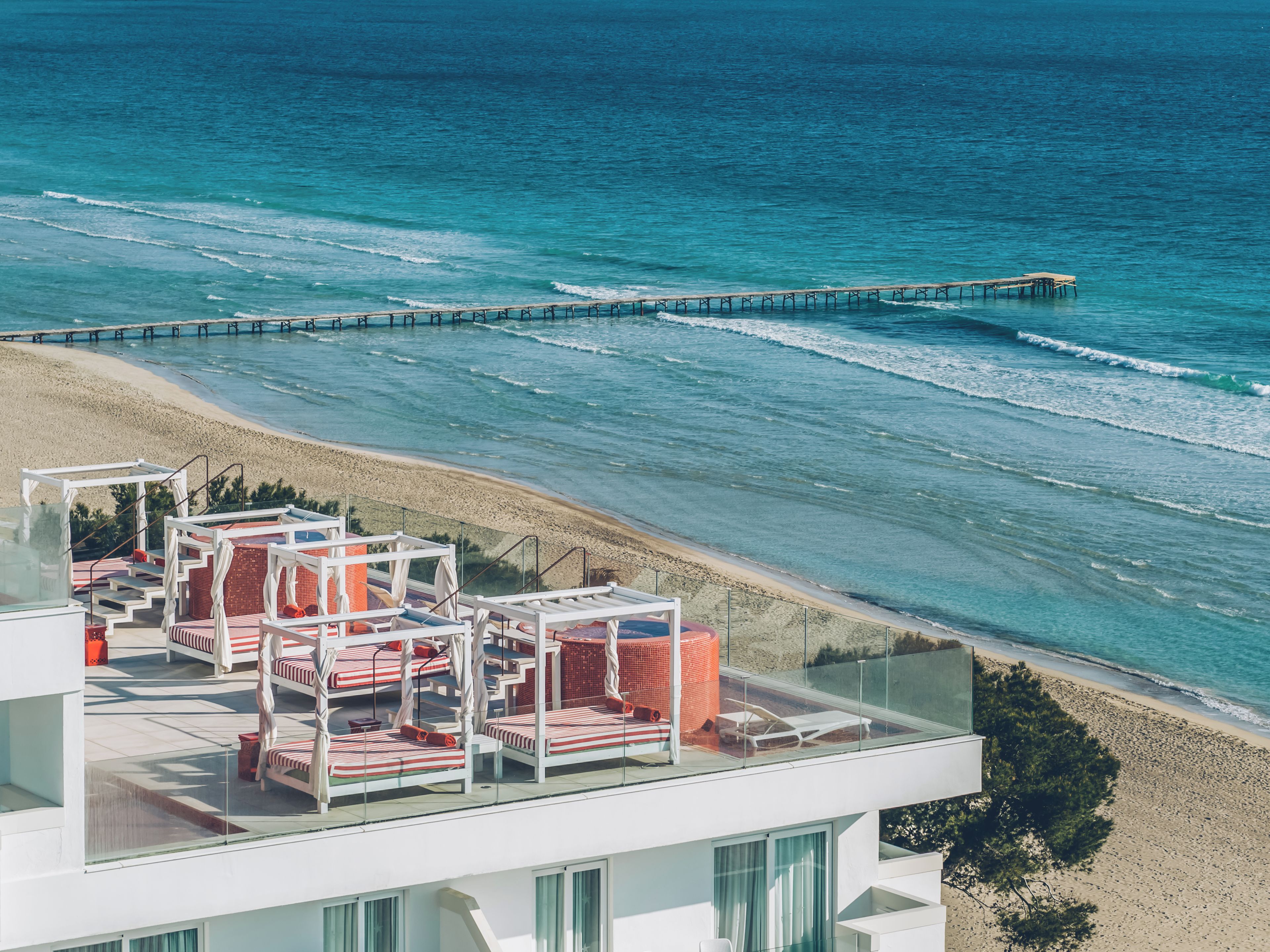 A beachfront hotel with a glass balustrade, red and white striped lounge chairs, and a pier extending into the sea.