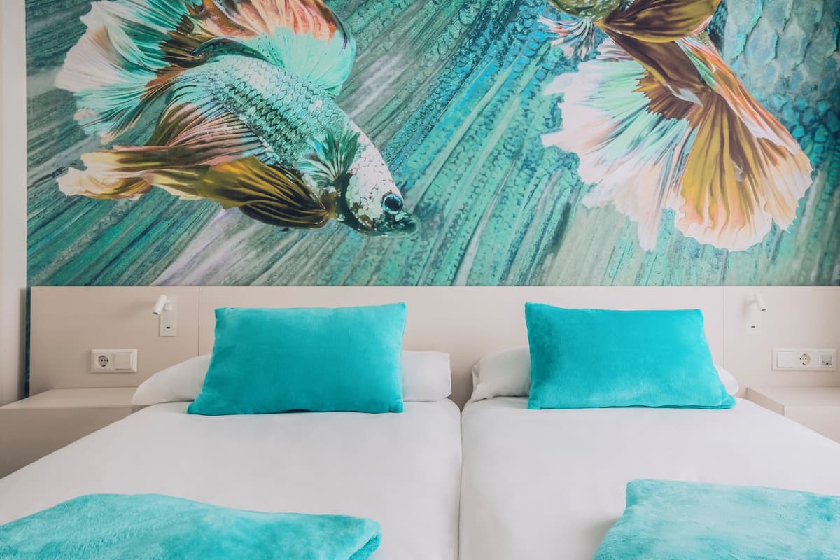 A hotel room with two beds, each with a white mattress and a teal pillow.