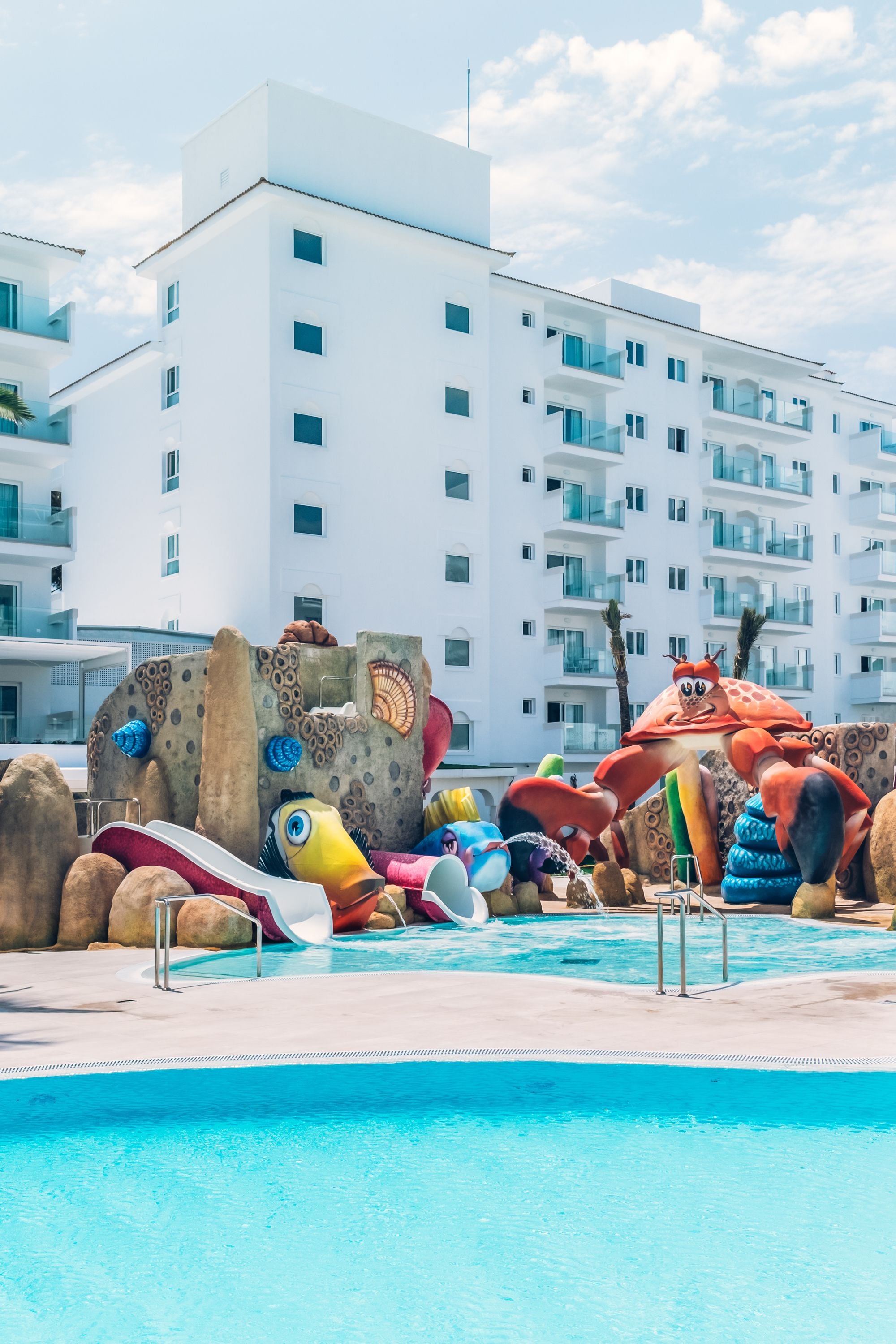 A swimming pool with inflatable sea creatures and a large crab statue, surrounded by a white building with balconies.