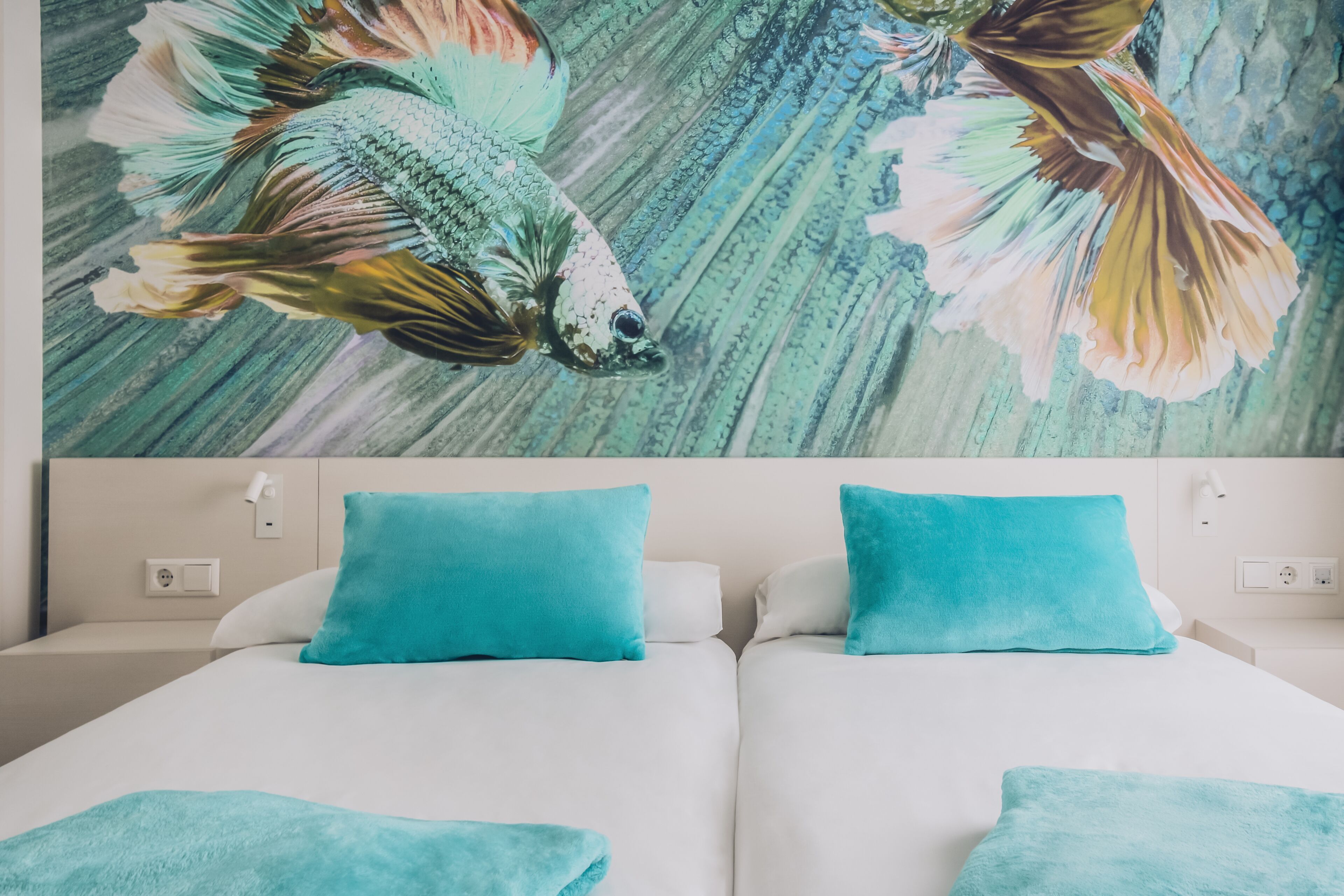 Une chambre d’hôtel avec une grande fresque représentant des poissons sur le mur, deux lits avec des coussins assortis de couleur turquoise et une tête de lit blanche.