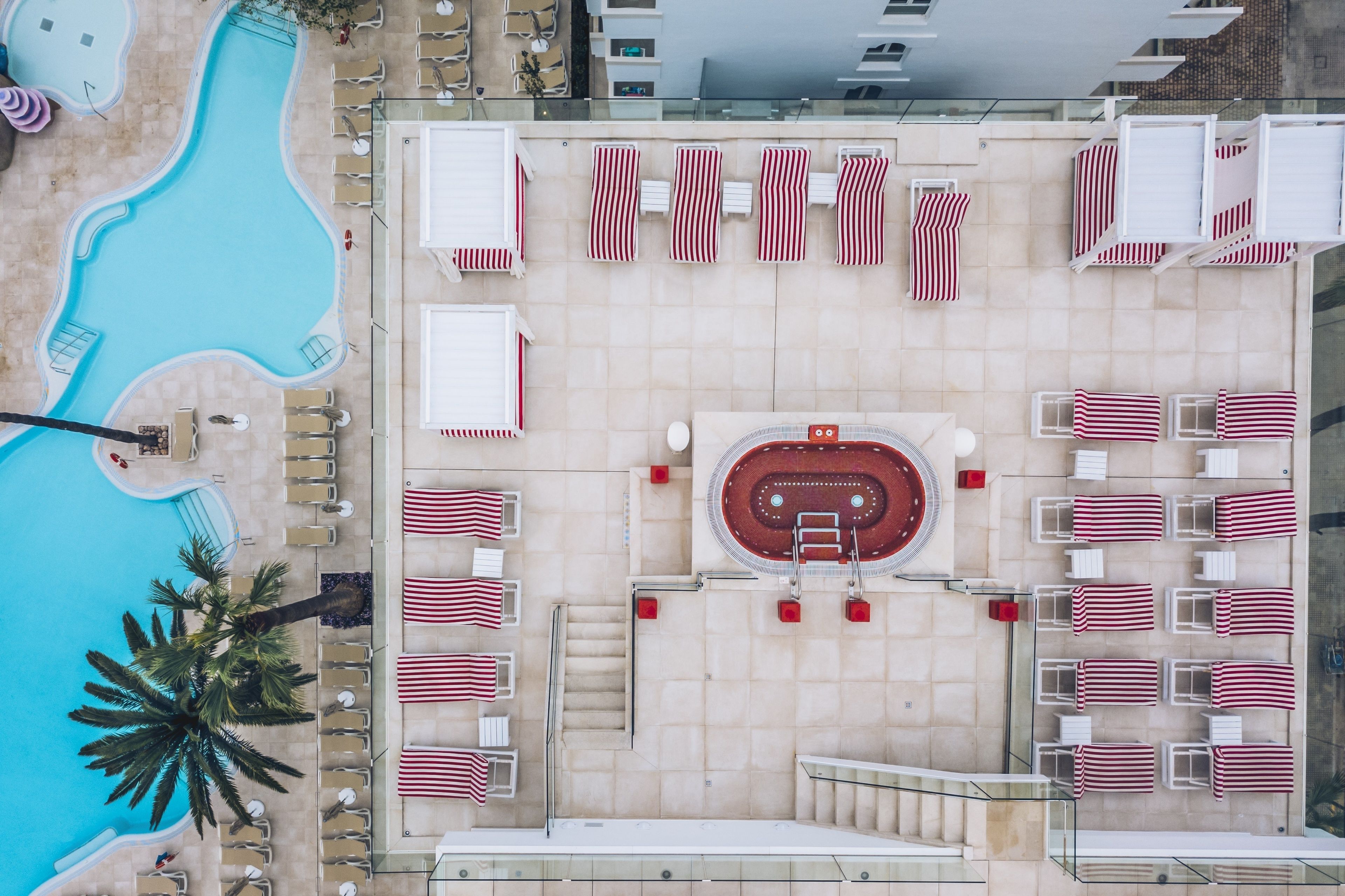 An aerial view of a hotel pool area with a hot tub, sun loungers, and a staircase leading to a terrace.