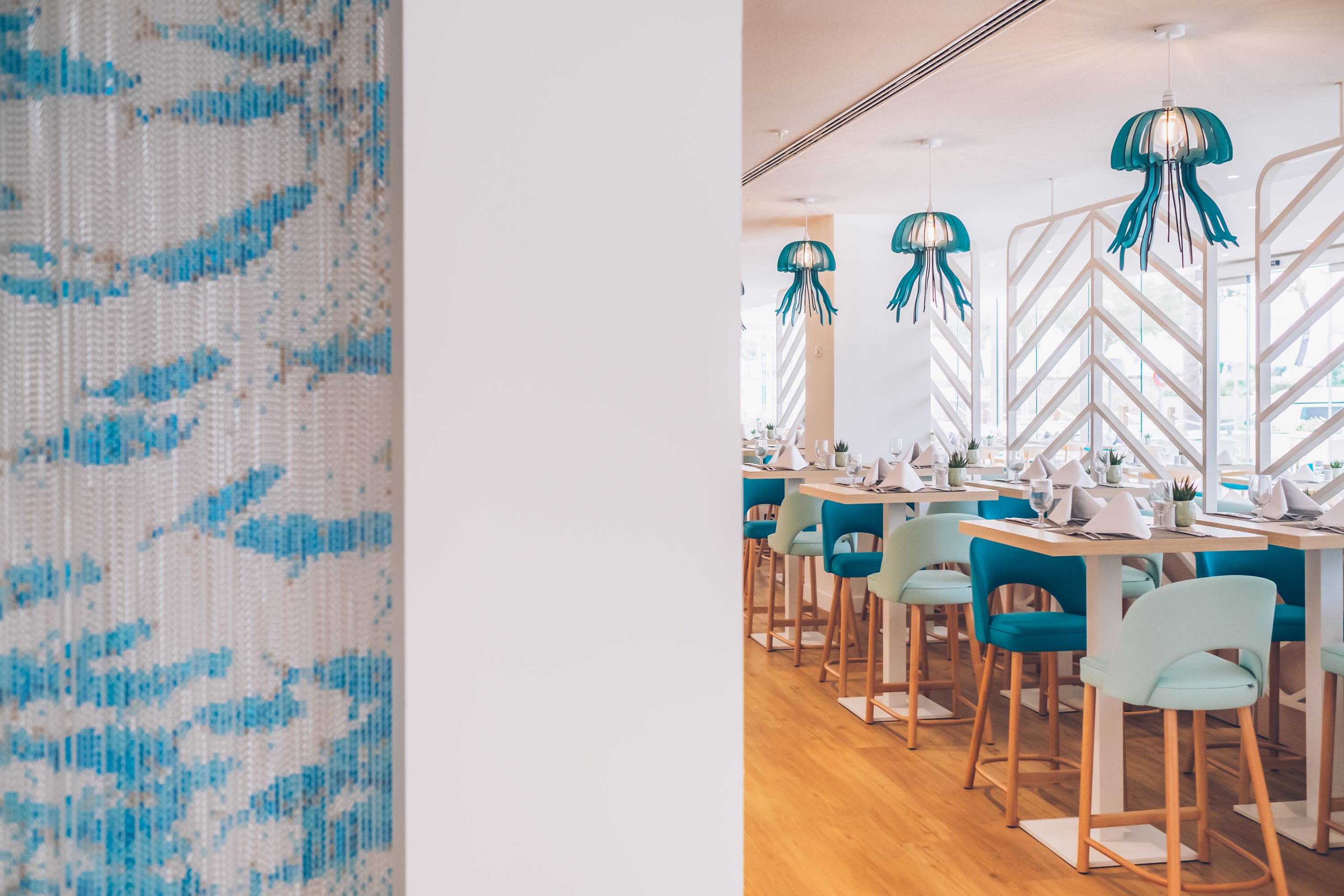 Un interior de restaurante con sillas color turquesa, mesas blancas y lámparas colgantes con forma de medusas color turquesa.