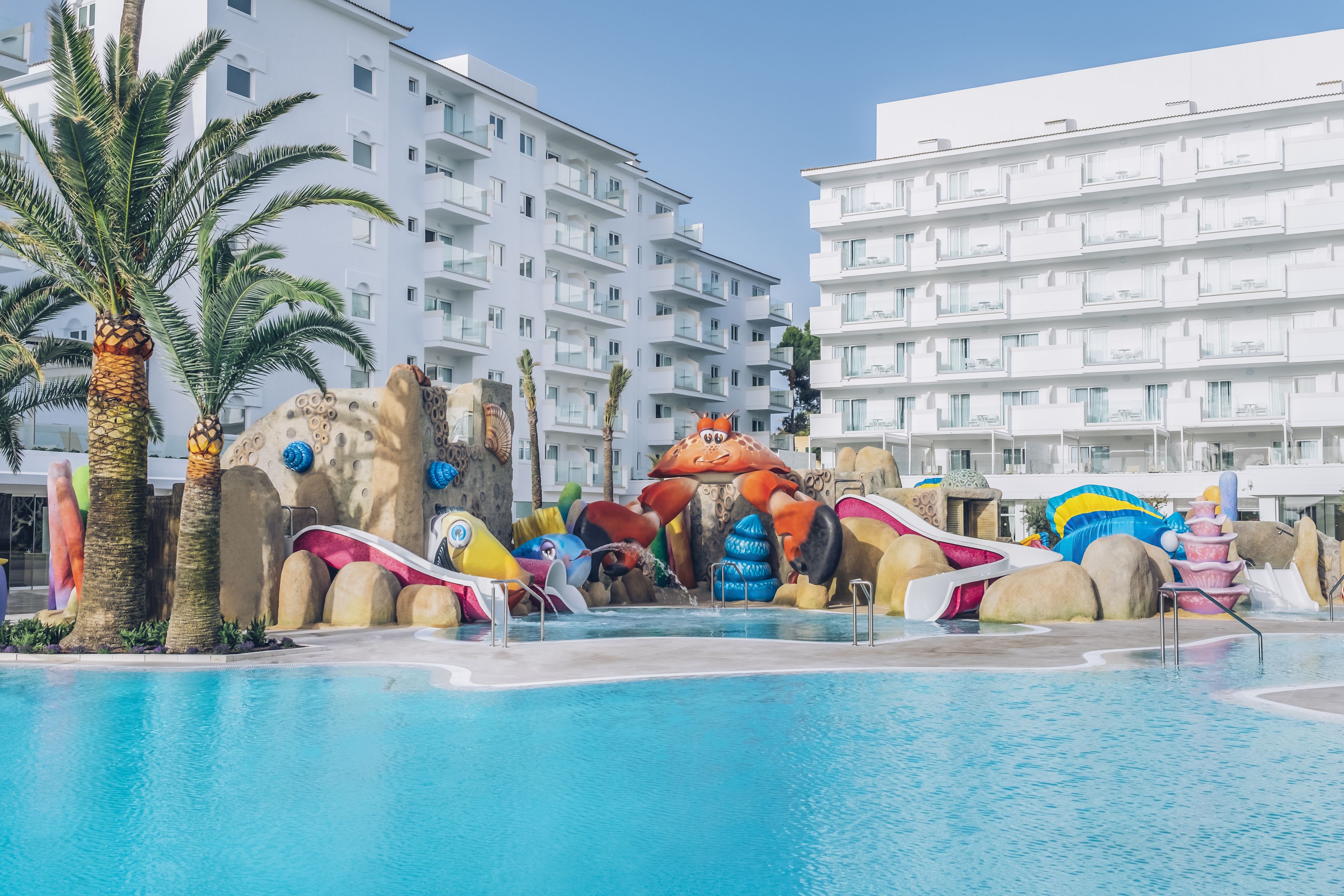 A swimming pool with slides and rock formations, surrounded by palm trees and white buildings.