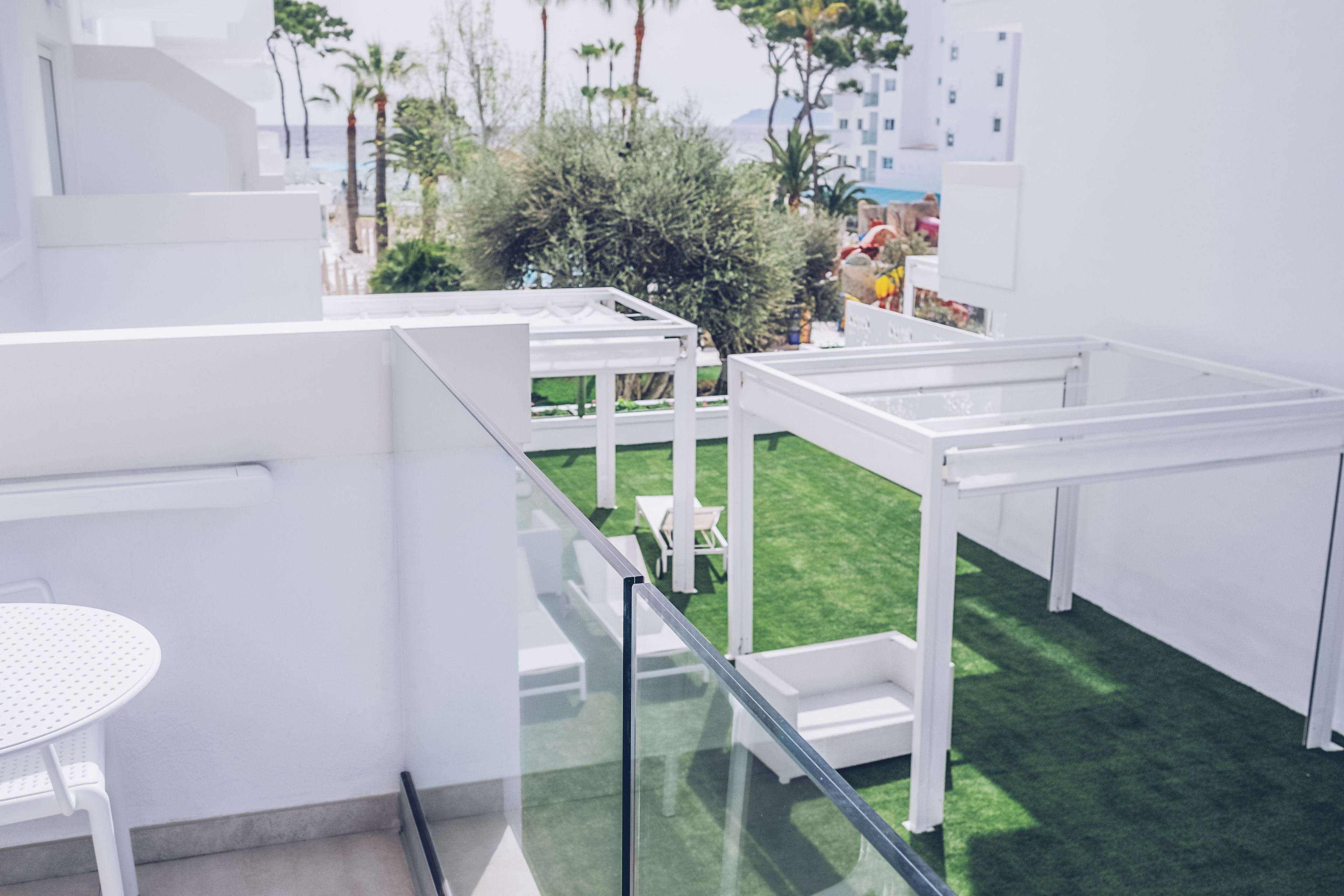Un espacio exterior moderno con mobiliario blanco, barandilla de vidrio y césped artificial verde.