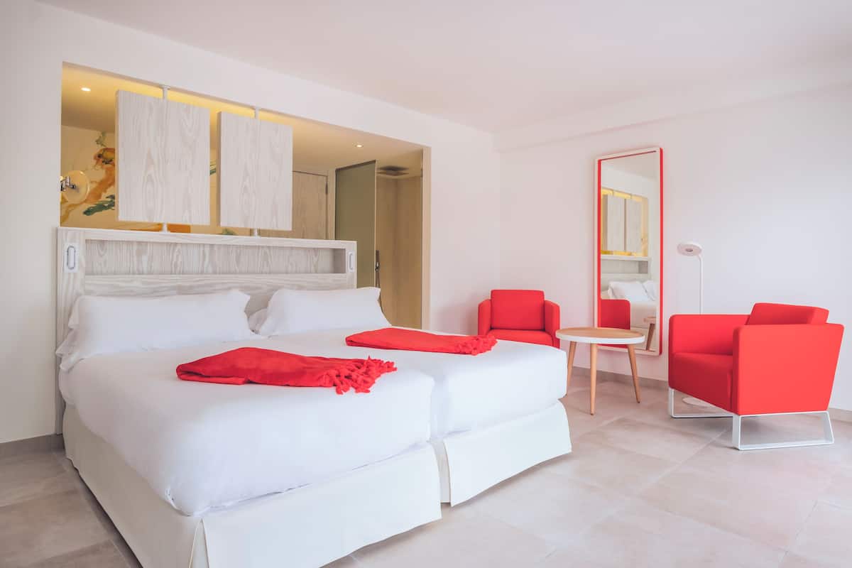 A modern hotel room with a large bed, two red chairs, a small table, and a mirror.