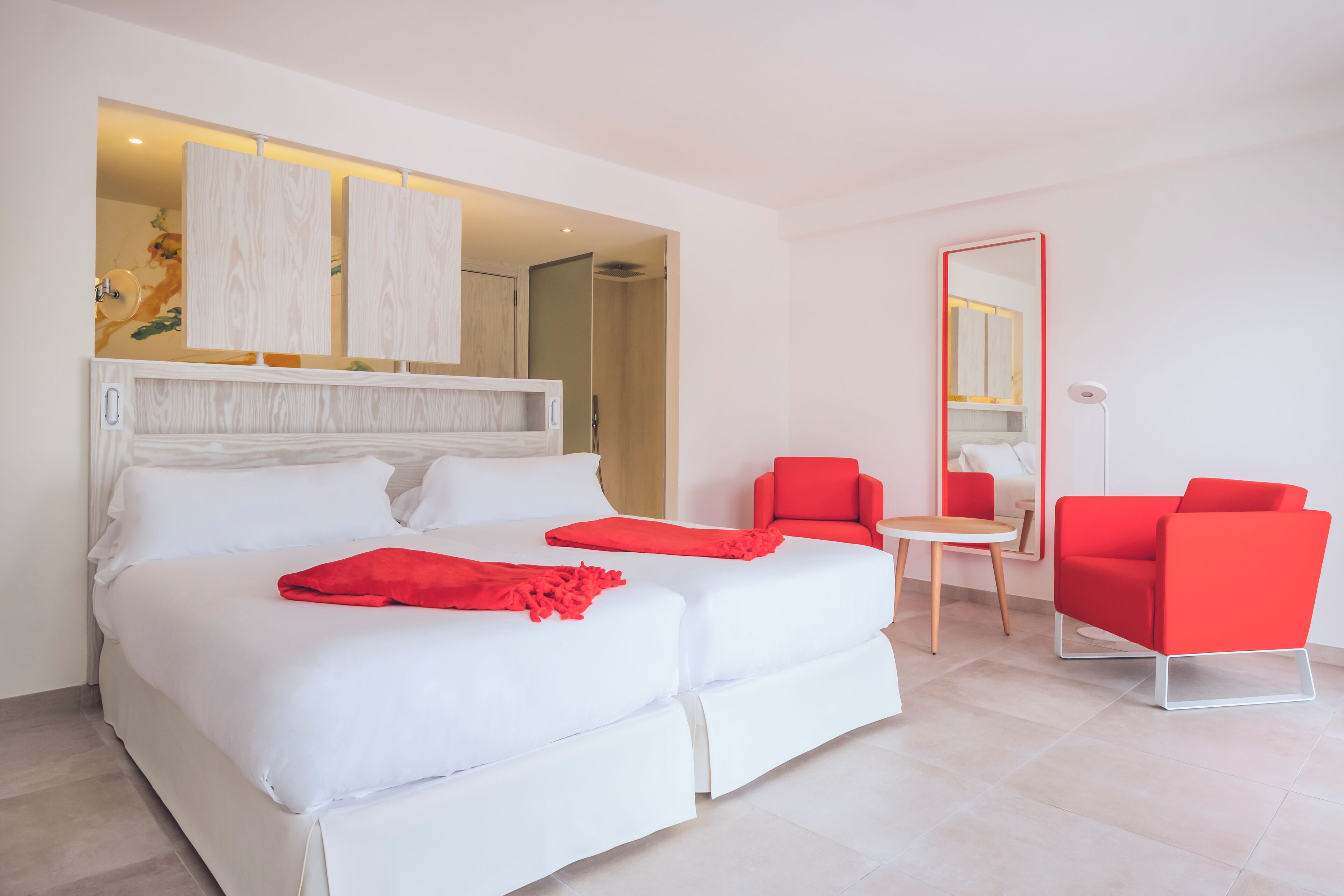 A modern hotel room with a large bed, two red chairs, a small table, and a mirror.