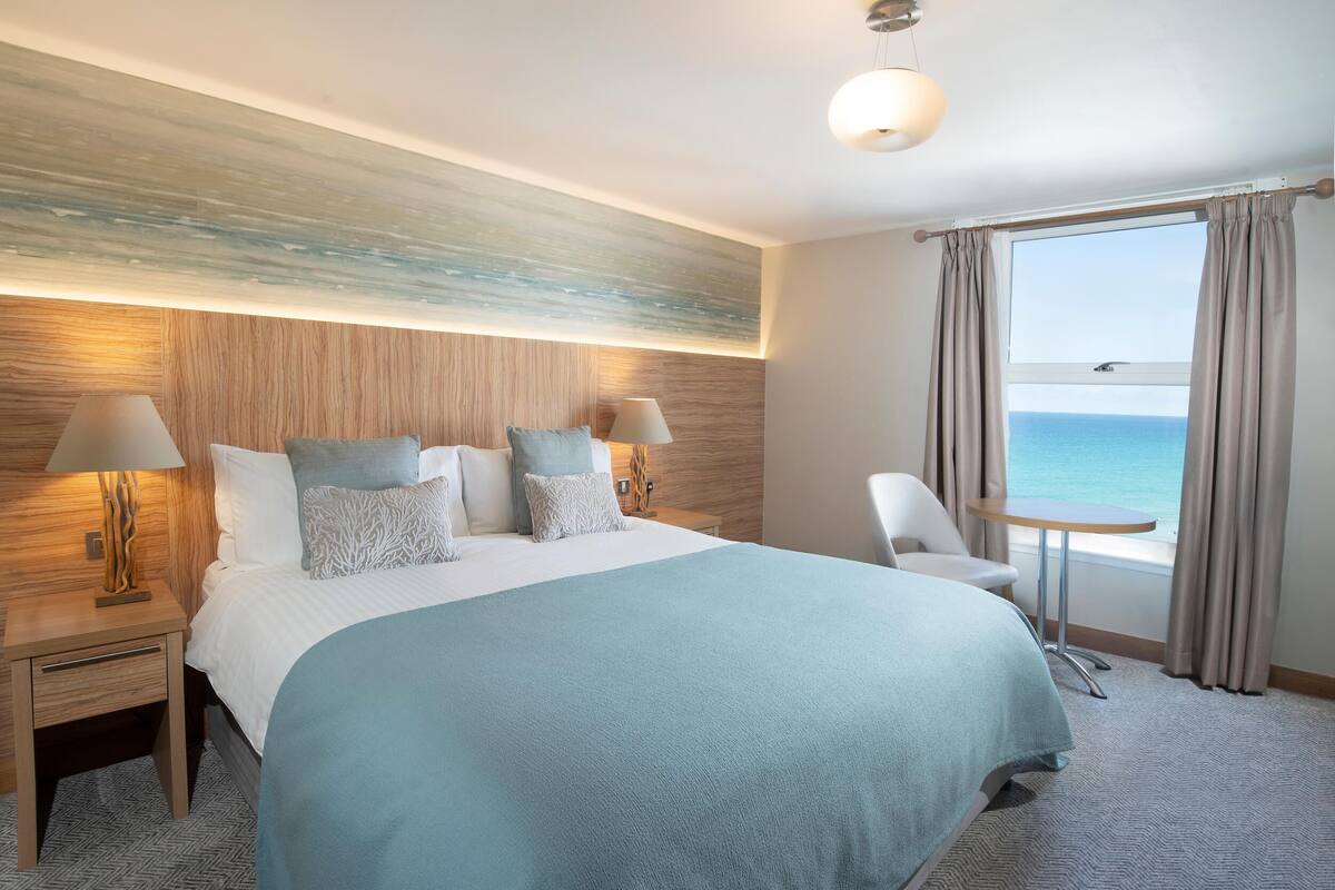 Een hotelkamer met een groot bed, twee nachtkastjes met lampen, een klein rond tafeltje, een stoel en uitzicht op de zee door een raam met gordijnen.