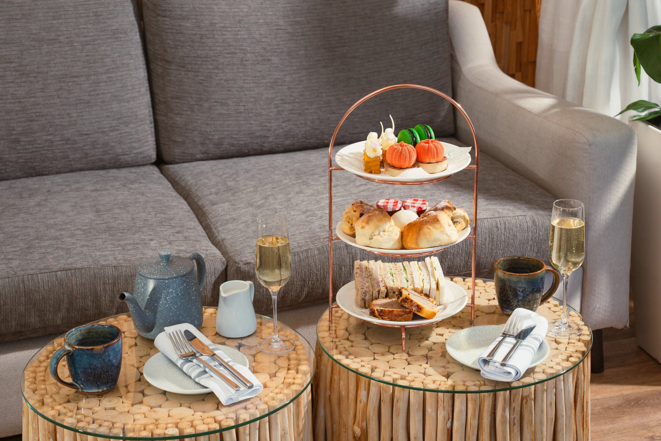 A three-tiered dessert stand with various pastries, a teapot, a glass of white wine, and a cup on a round wooden table.