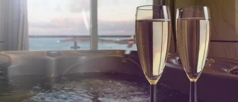 Dos copas de champán sobre una mesa, con un jacuzzi al fondo y vista al mar a través de una ventana.