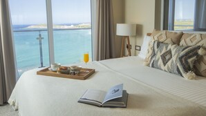 Une chambre d’hôtel avec un grand lit, un plateau avec des articles pour le petit-déjeuner, un livre et une vue sur la mer par de grandes fenêtres.