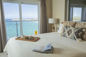 Une chambre d’hôtel avec un grand lit, un plateau avec des articles pour le petit-déjeuner, un livre et une vue sur la mer par de grandes fenêtres.
