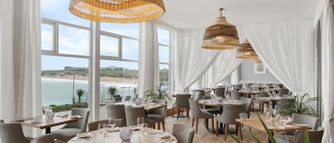 Un restaurante con mesas redondas, cortinas blancas y lámparas colgantes de mimbre.