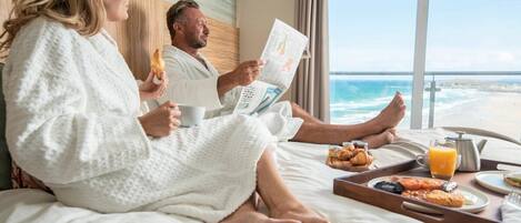A couple in a hotel room with a bed, breakfast tray, and a view of the beach.