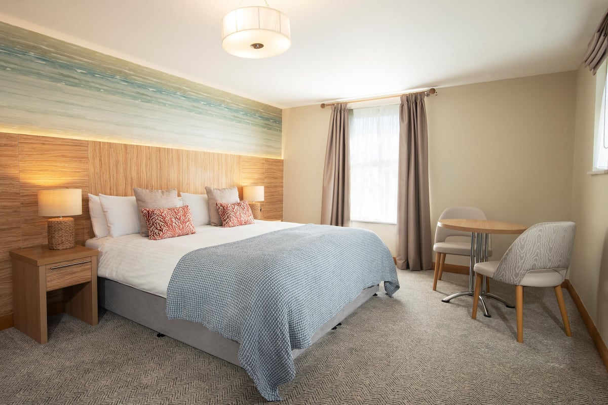 Een hotelkamer met een groot bed, nachtkastjes met lampen, een klein tafeltje met stoelen, en uitzicht op het omliggende landschap door een raam.