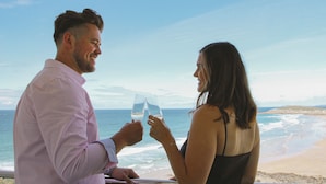Un couple trinque avec des verres de vin dans un cadre idyllique, au bord de la plage, avec vue sur l’océan.