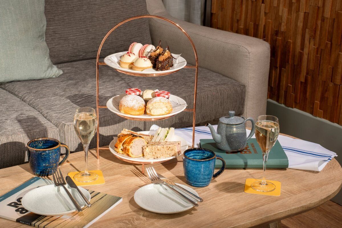A three-tiered dessert stand on a wooden table with plates of pastries, a teapot, and two glasses of a clear liquid, likely wine, in a room with a grey sofa and a wooden wall.