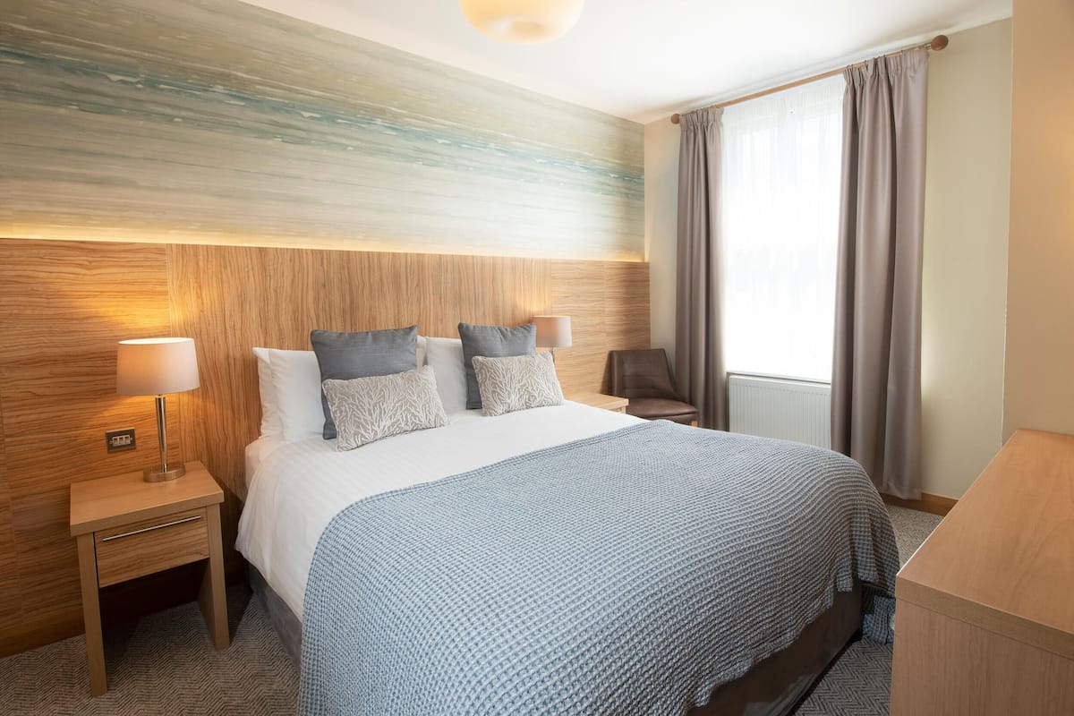 Een hotelkamer met een groot bed, twee nachtkastjes met lampen, een houten hoofdeinde en uitzicht op het omliggende landschap door een raam met gordijnen.