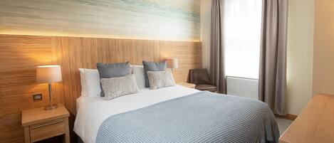 Habitación de hotel con una cama grande, dos mesitas de noche con lámparas, un cabecero de madera y vistas al exterior a través de una ventana con cortinas.