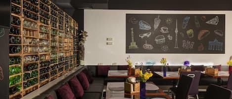 A restaurant with a wall of green glassware, a chalkboard with food illustrations, and purple seating.