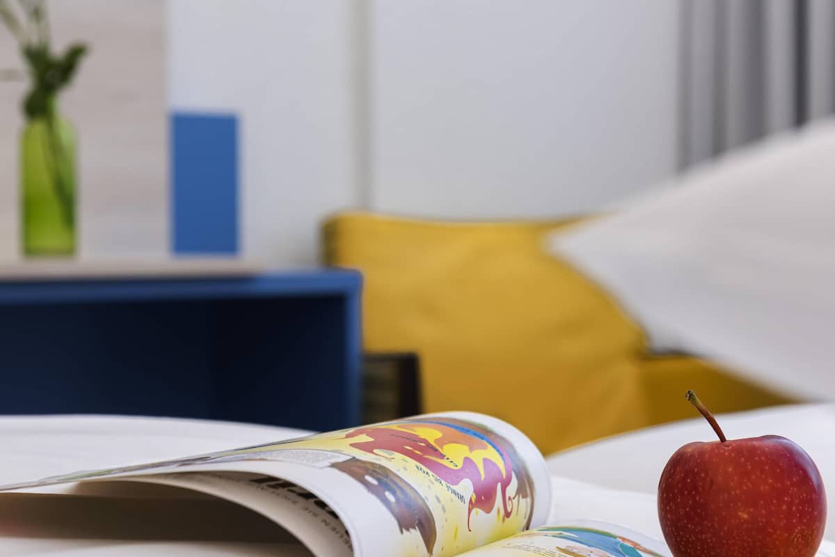 Uma revista com capa colorida, uma maçã vermelha, uma cadeira azul e um travesseiro amarelo em um quarto.