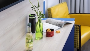 A table with a bottle, a green vase with a flower, a black tumbler, a red cup, and a magazine. - St Christopher's Hotel Berlin - Alexanderplatz (Berlin)