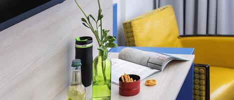 A table with a bottle, a green vase with a flower, a black tumbler, a red cup, and a magazine.