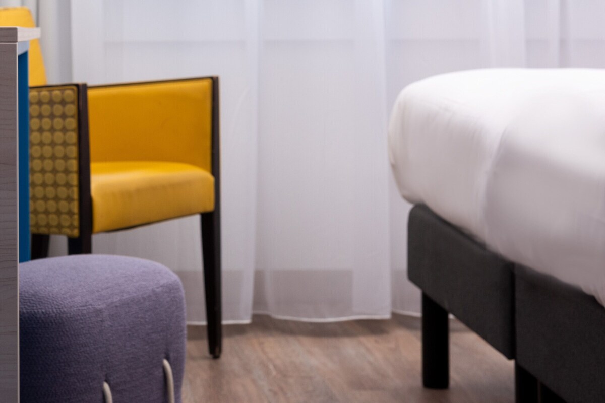 A hotel room with a bed, a chair, and a desk with a lamp.
