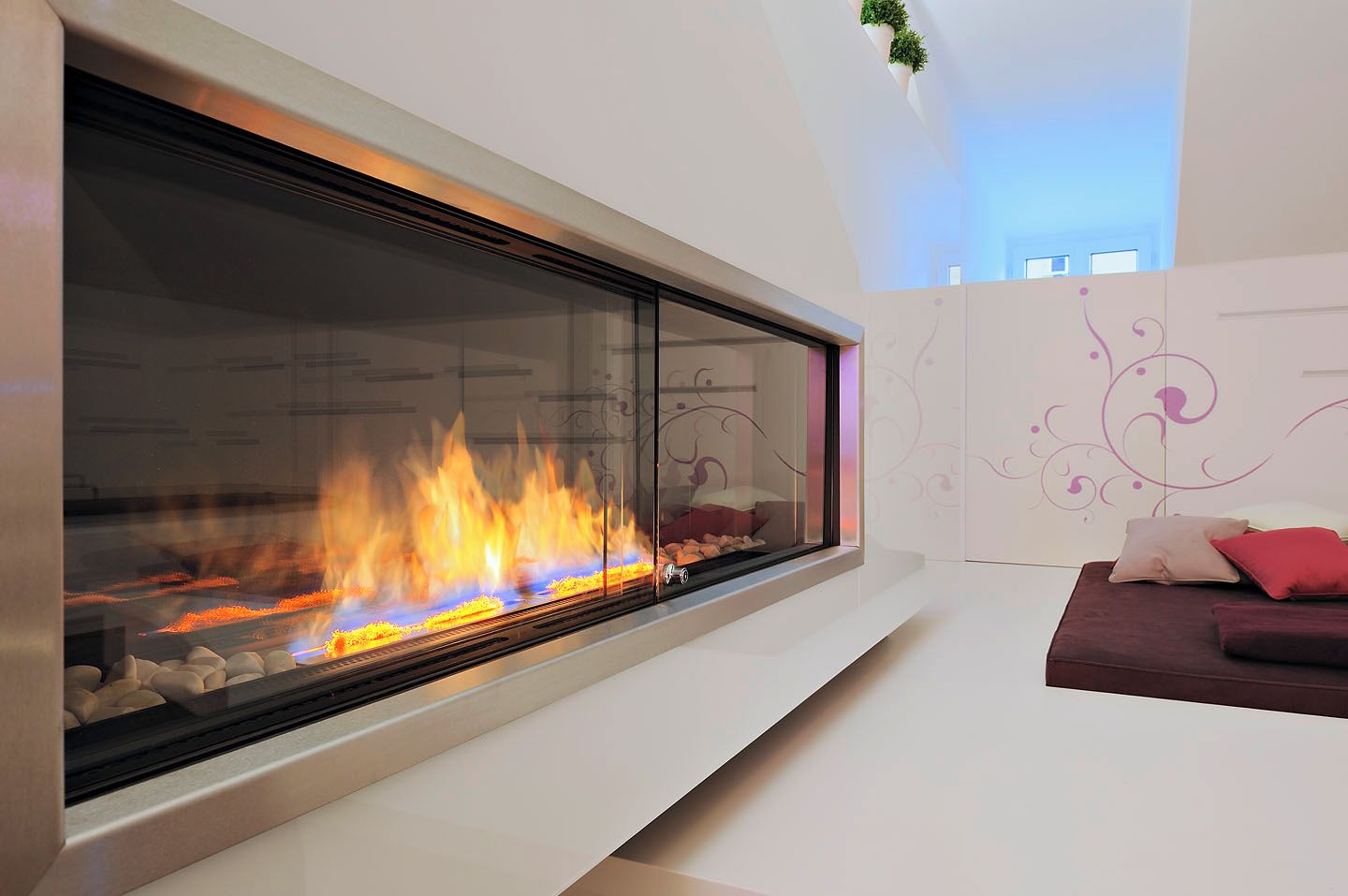 A modern living room with a large glass fireplace, a white wall with decorative patterns, and a cushioned seating area.