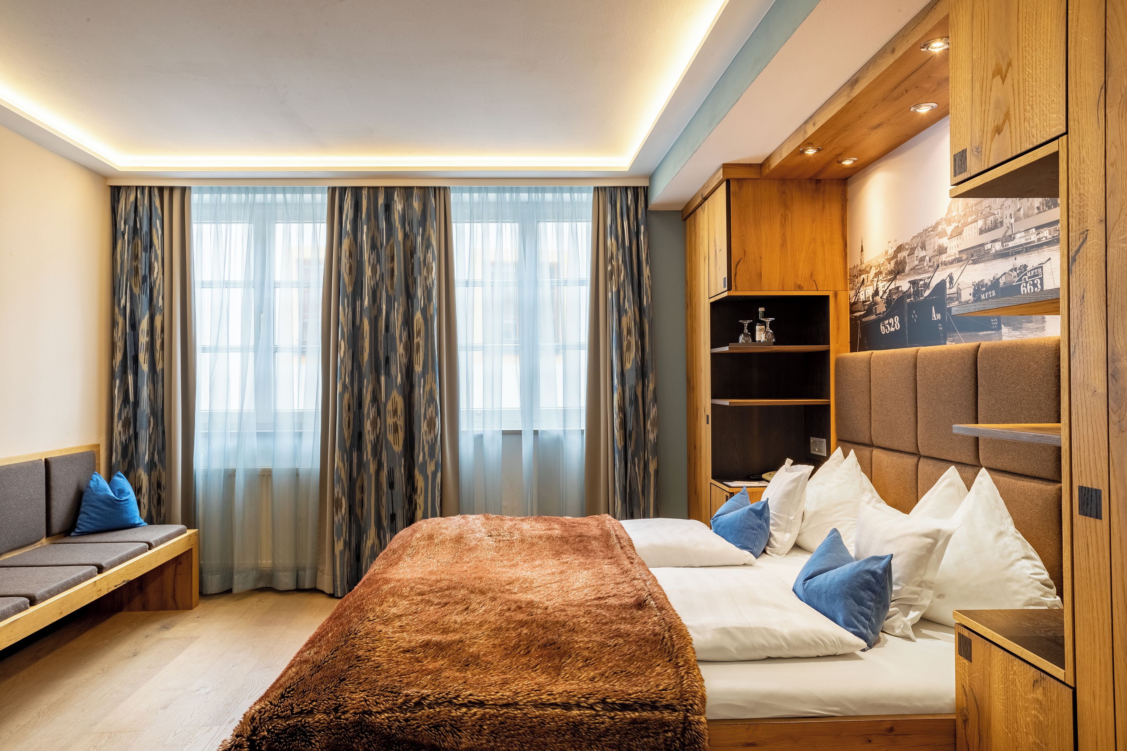 Una habitación de hotel con cama, una zona de estar, un armario de madera y un ventanal con cortinas.