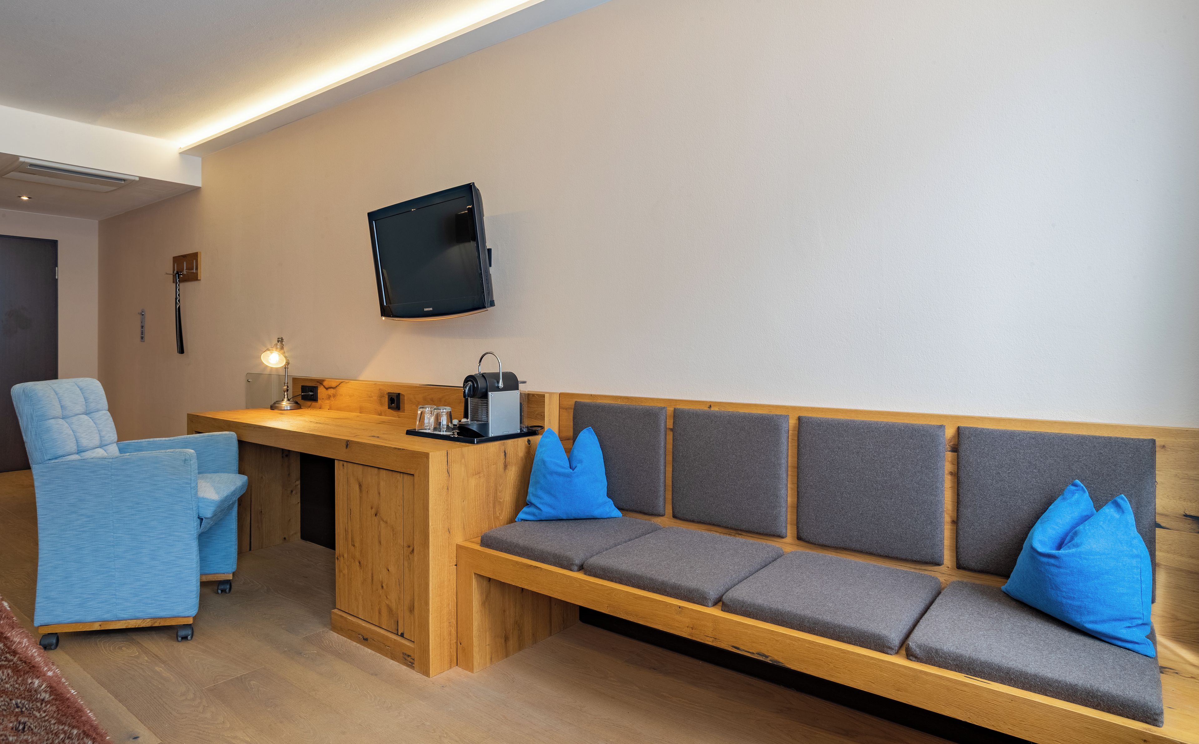 A room with a wooden bench, a blue chair, a flat-screen TV mounted on the wall, and a coffee machine on the bench.