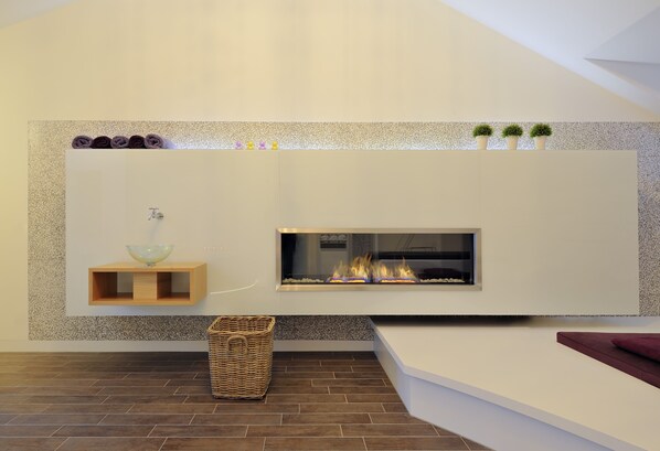 A modern living room with a built-in fireplace, a wooden shelf with towels, and a wicker basket on the floor. - Hotel Residenz Passau (Passau)