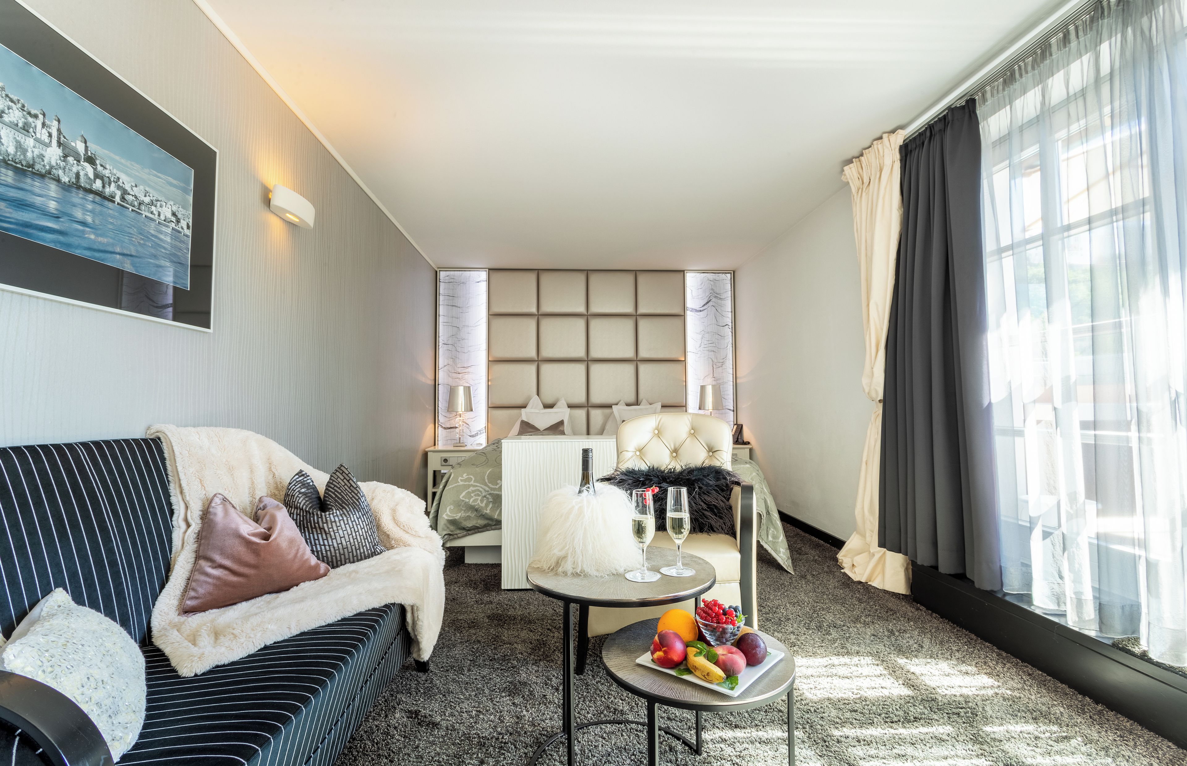A modern hotel room with a sofa, a small round table with fruit and drinks, a bed with a headboard, and a large window with curtains.
