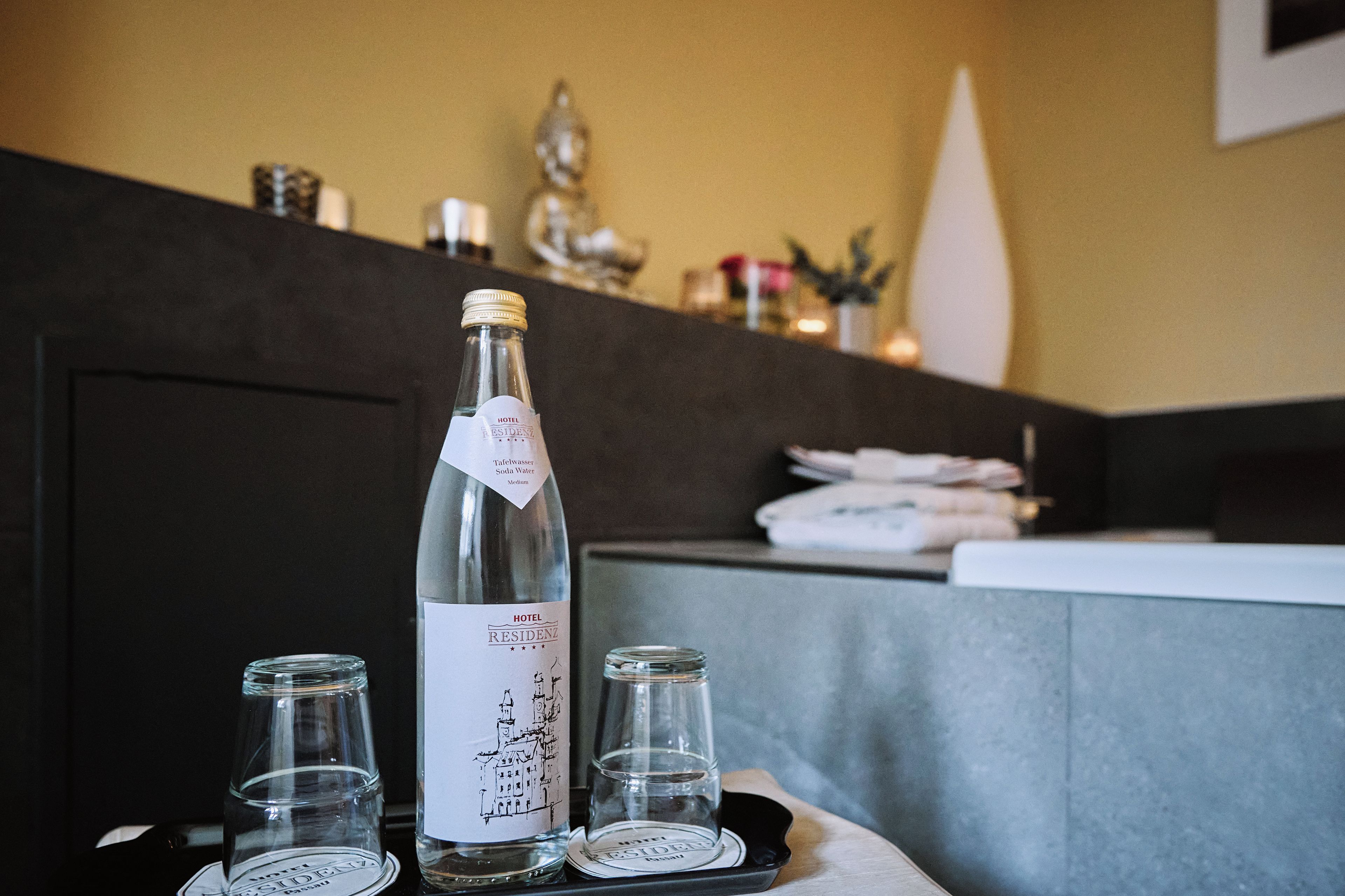 Une bouteille d’eau de la marque Hotel Residency, avec une étiquette, deux verres transparents et un dessous de verre, le tout posé sur une table.