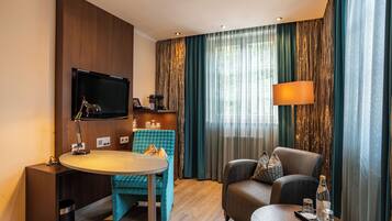 A hotel room with a flat-screen TV mounted on the wall, a round dining table with a chair, a sofa, and a small side table with a bottle and glasses.