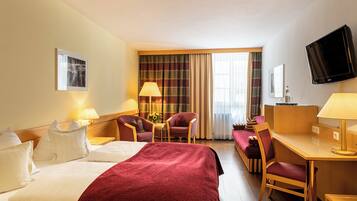 Een hotelkamer met een bed, twee fauteuils, een bureau, een televisie en een raam met gordijnen.