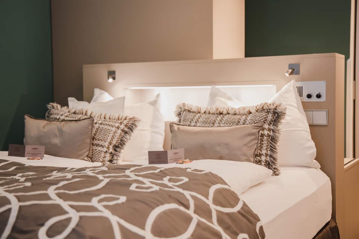 Ein Hotelzimmer mit einem großen Bett, zahlreichen Kissen, einem Nachttisch mit Lampe sowie einer wandmontierten Bedieneinheit.