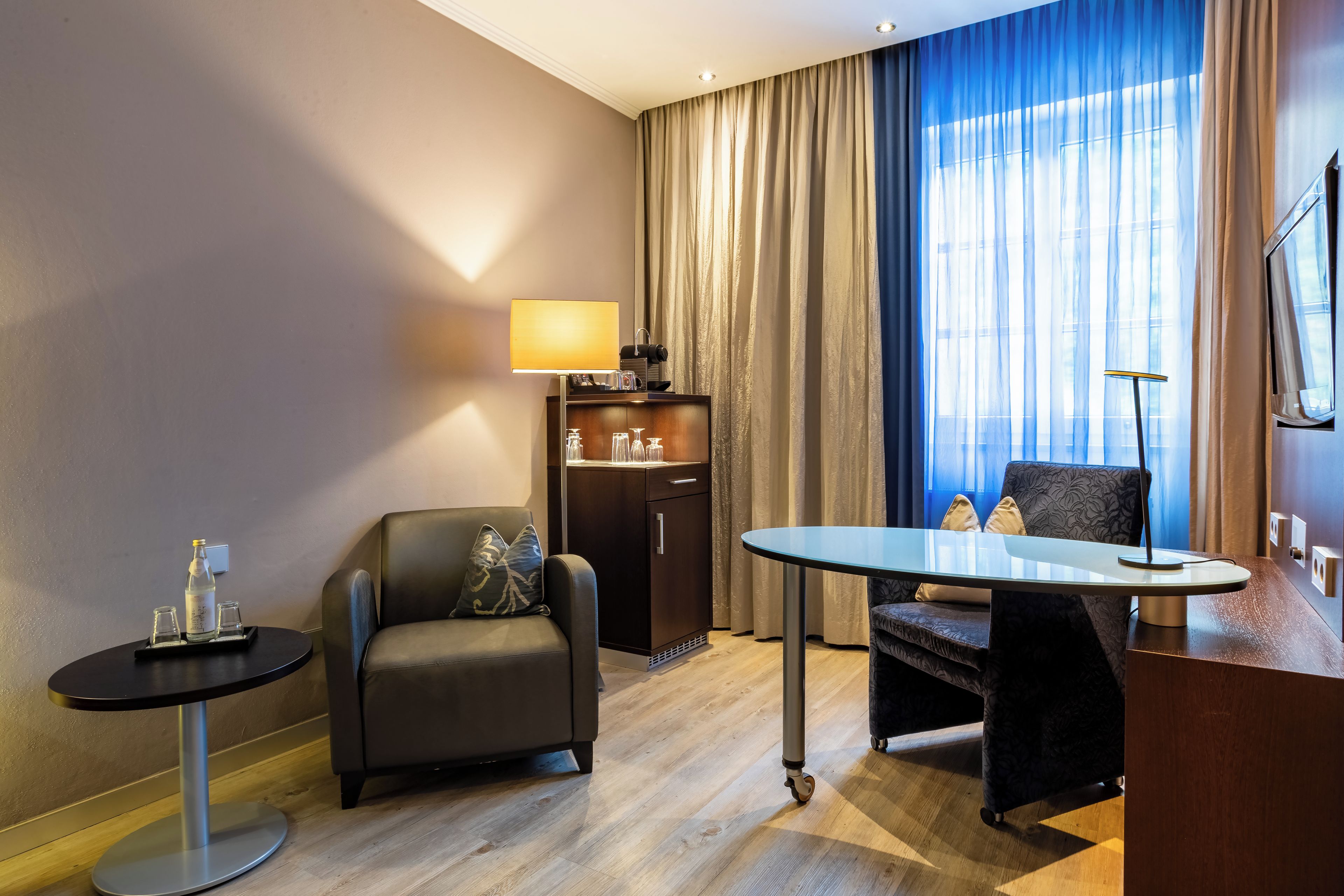 Ein Hotelzimmer mit einer Couch, einem kleinen Tisch, einem Schrank und einem großen Fenster mit Vorhängen.
