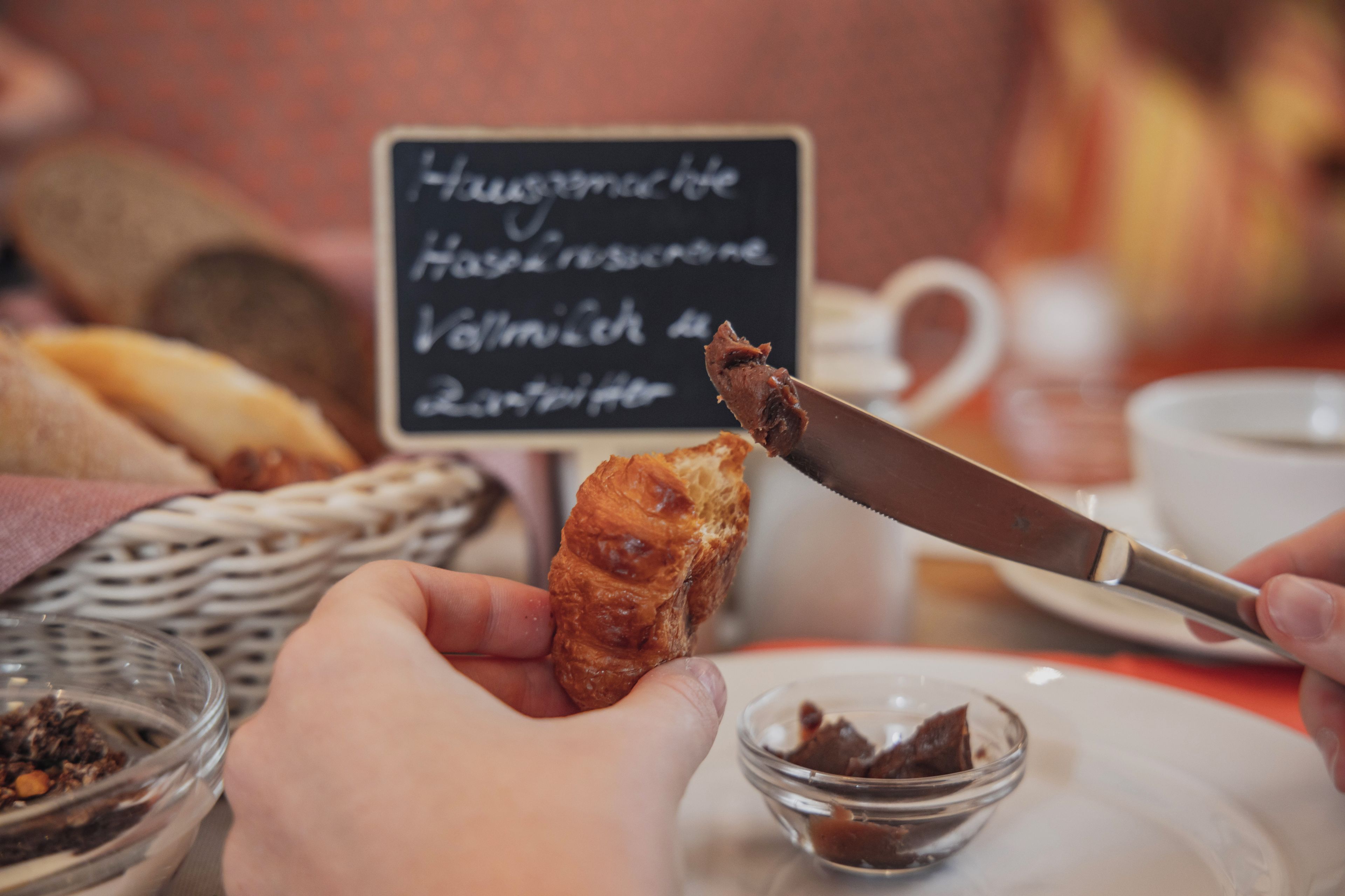 A person holding a pastry with a knife, a bowl of chocolate spread, and a sign with text in the background.