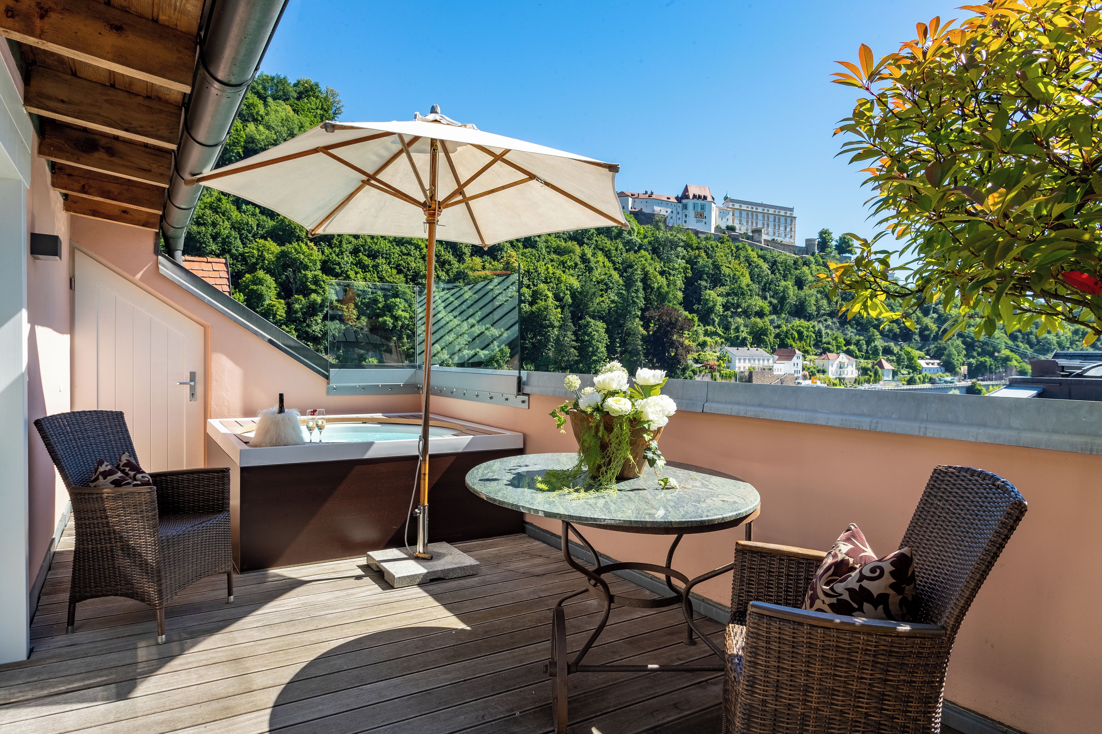Eine Dachterrasse mit Whirlpool, zwei Korbstühlen, einem Tisch mit Blumenvase und einem großen Sonnenschirm.
