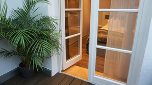 Una habitación con piso de madera, una cama con cabecera blanca y una planta grande en maceta cerca de la puerta.