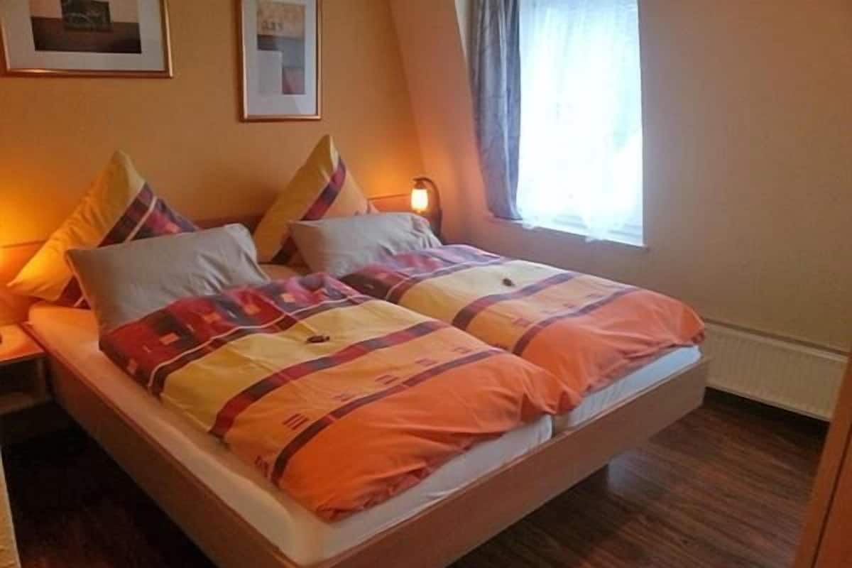 A double bed with a striped blanket and multiple pillows, a bedside table, and a window with curtains.
