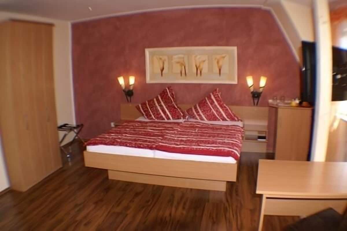 A bedroom with a bed, wooden floor, and a red wall with a framed artwork.