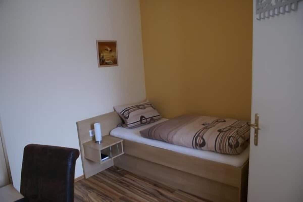 A small, single-bed room with a wooden floor, a bedside table with a lamp, and a chair.