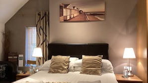 A hotel room with a large bed, two bedside tables with lamps, a painting of a beach scene, and a window with blinds. - The Manian (Kilgetty)