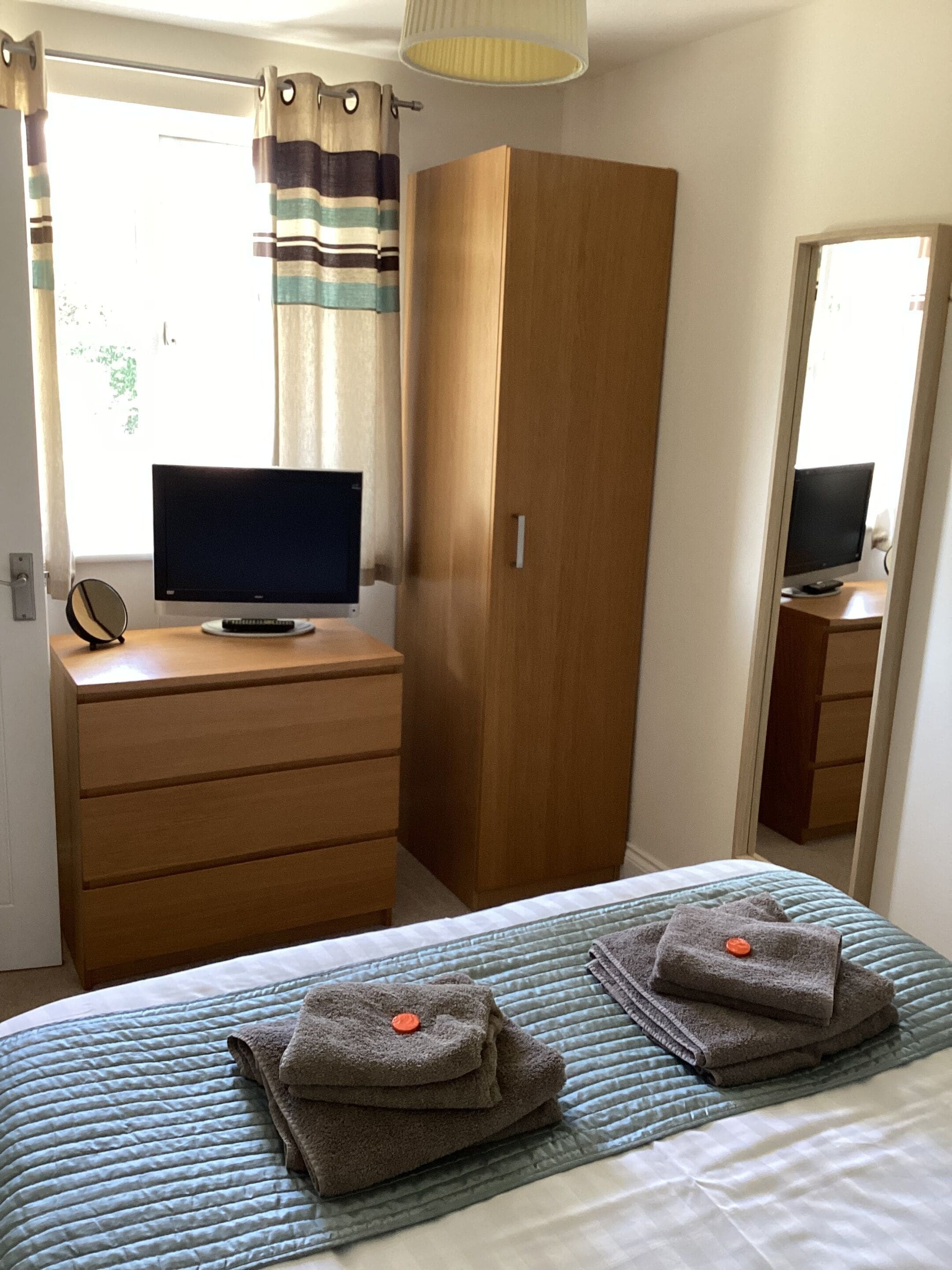 A bedroom with a bed, a wooden wardrobe, a desk with a computer, and a mirror.